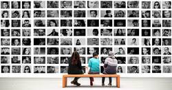 Drei Personen sitzen auf einer Bank und betrachten eine Wand voller Bilder (Symbolfoto Ausstellung)