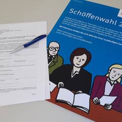 Der Bewerbungsbogen für die Jugendschöffenwahl im Saale-Orla-Kreis liegt neben dem blauen Plakat des Thüringer Justizministeriums zur Schöffenwahl 2023 auf einem Tisch (Symbolfoto).