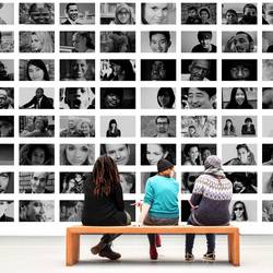 Drei Personen sitzen auf einer Bank und betrachten eine Wand voller Bilder (Symbolfoto Ausstellung)